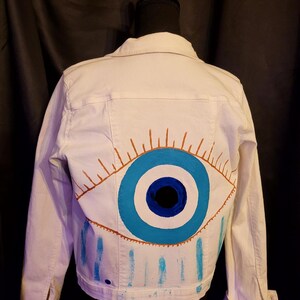 Può includere: Giacca di jeans bianca con un grande disegno di occhio dipinto a mano sul retro. L'occhio presenta dettagli blu e marroni, con gocce di vernice blu. Un capo di abbigliamento esterno classico e casual.