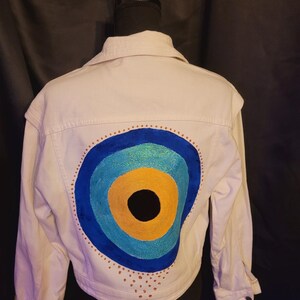 Könnte beinhalten: Weiße Jeansjacke mit einem großen, farbenfrohen Augendesign auf dem Rücken. Das Auge hat konzentrische Kreise in Blau, Türkis, Gelb und einen schwarzen Mittelpunkt. Kleine goldene Punkte umgeben das Auge.