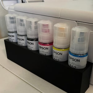 May include: A white Epson printer with a black ink cartridge holder. The holder contains six ink bottles in various colors: black, pink, yellow, and blue. The ink bottles are labeled with the Epson brand and the number 552.
