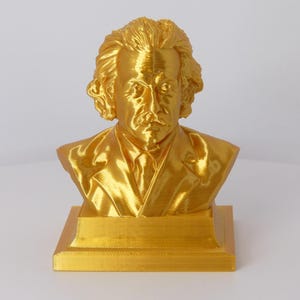 May include: A metallic gold-colored bust sculpture of Albert Einstein, featuring his signature wild hair and mustache. The figure is depicted wearing a suit jacket and tie, mounted on a square base, capturing the likeness of the physicist.