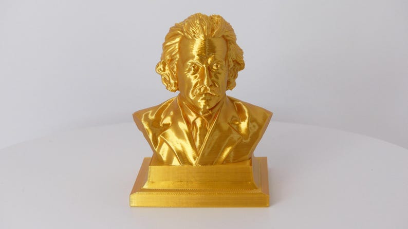 May include: A metallic gold-colored bust sculpture of Albert Einstein, featuring his signature wild hair and mustache. The figure is depicted wearing a suit jacket and tie, mounted on a square base, capturing the likeness of the physicist.