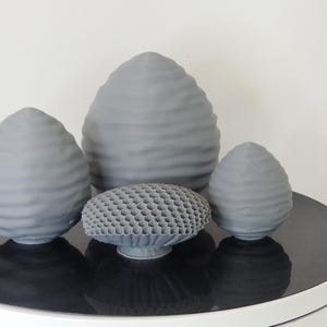 Puede incluir: Cuatro esculturas de cerámica gris sobre una superficie negra. Tres tienen forma de huevo con una superficie texturizada. La cuarta es una escultura redonda con forma de panal.