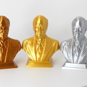 Könnte beinhalten: Drei 3D-gedruckte Büsten einer Person in verschiedenen Metallic-Oberflächen: Gold, Messing und Silber. Die Büsten stehen auf kleinen quadratischen Sockeln.