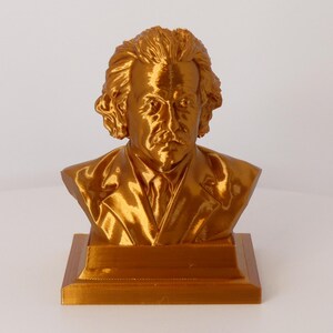 May include: A metallic gold-colored bust of Albert Einstein featuring his signature wild hair, mustache, and a suit jacket. The sculpture is mounted on a square base and displays a reflective, glossy finish across the entire surface of the figure.