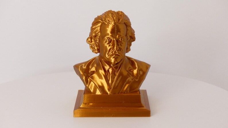 May include: A metallic gold-colored bust of Albert Einstein featuring his signature wild hair, mustache, and a suit jacket. The sculpture is mounted on a square base and displays a reflective, glossy finish across the entire surface of the figure.
