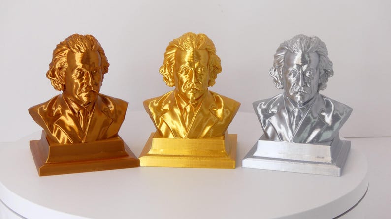May include: Three busts of Albert Einstein arranged in a row, each featuring a distinct metallic finish. The left bust is bronze, the center bust is gold, and the right bust is silver. Each figure is mounted on a square base with a smooth surface.
