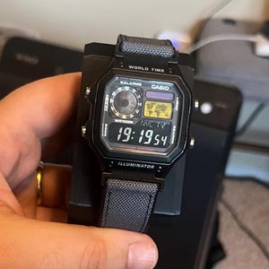 May include: Black Casio digital watch with a gray fabric band. The watch face displays the time, date, and other functions. The watch is on a black stand. The text "CASIO" and "ILLUMINATOR" are visible.