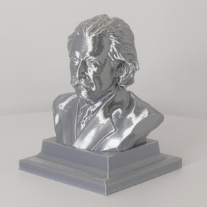 May include: A metallic silver-colored bust of Albert Einstein featuring his signature wild hair and mustache. The sculpture is mounted on a solid, square-shaped base with a stepped design, showcasing a smooth, reflective finish throughout the piece.