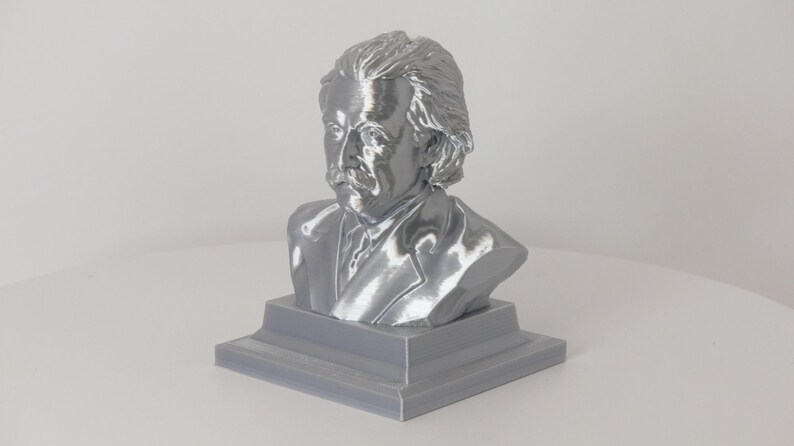 May include: A metallic silver-colored bust of Albert Einstein featuring his signature wild hair and mustache. The sculpture is mounted on a solid, square-shaped base with a stepped design, showcasing a smooth, reflective finish throughout the piece.