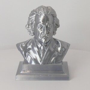 May include: A metallic silver-colored bust of Albert Einstein featuring his signature wild hair, mustache, and suit jacket. The sculpture is mounted on a square base and displays a smooth, reflective finish that captures light across its detailed features.