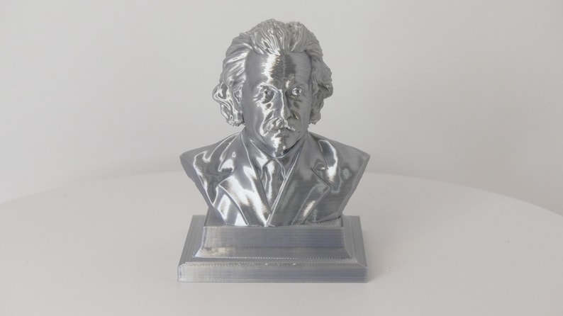 May include: A metallic silver-colored bust of Albert Einstein featuring his signature wild hair, mustache, and suit jacket. The sculpture is mounted on a square base and displays a smooth, reflective finish that captures light across its detailed features.