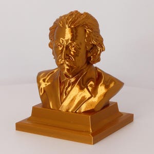 May include: A metallic gold-colored bust of Albert Einstein featuring his signature wild hair and mustache. The sculpture is mounted on a square, tiered base and displays a smooth, reflective finish that captures light across the facial features.