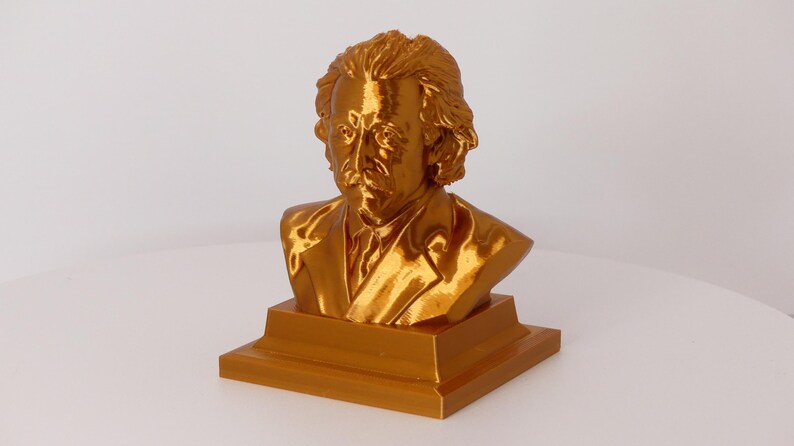May include: A metallic gold-colored bust of Albert Einstein featuring his signature wild hair and mustache. The sculpture is mounted on a square, tiered base and displays a smooth, reflective finish that captures light across the facial features.