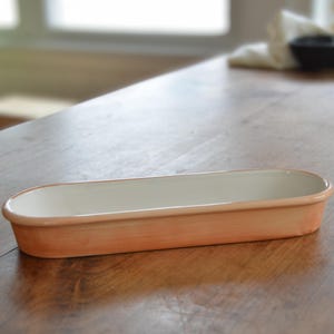 May include: A rectangular ceramic baking dish with a light peach exterior and a white interior. The dish has rounded edges and is set on a wooden surface. The dish is approximately 12 inches long.