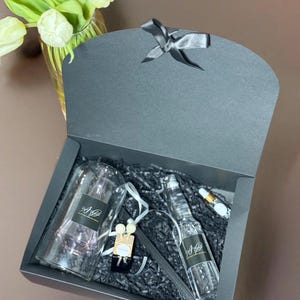 Op de afbeelding: Een zwarte cadeaudoos met een zwart lint, gevuld met een doorzichtige glazen fles met een zwart etiket, een doorzichtige glazen diffuser met roze vloeistof en een kleine set houten diffuserstokjes. De doos is gevuld met zwart geribbeld papier.