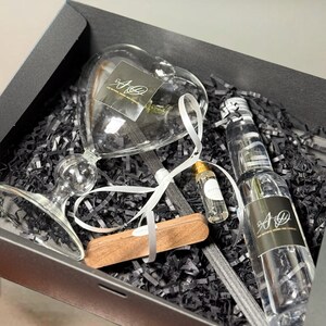 Könnte beinhalten: Eine schwarze Geschenkbox gefüllt mit schwarzem Schredderpapier. Die Box enthält einen herzförmigen Glasdiffusor, eine Flasche Diffusionsöl, einen Holzdiffusionsstab und eine kleine Flasche ätherisches Öl. Die Diffusionsölflasche trägt ein Etikett mit der Aufschrift "All Natural Home and Candles".