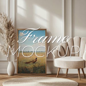May include: A framed print of a pheasant in a field, with the text "Frame Mockup" overlayed on the image. The print is hanging on a wall in a room with a white armchair and a vase of dried pampas grass.