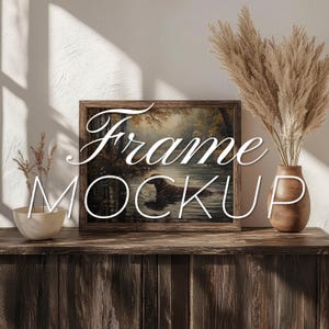 Puede incluir: Una maqueta de marco de madera con una pintura de una escena de bosque y un río. El marco está sentado en una mesa de madera con un jarrón de hierba de la pampa seca.