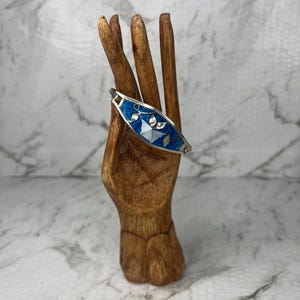 May include: A silver bracelet with a blue enamel inlay and inlaid mother-of-pearl and gold-colored accents. The bracelet is displayed on a wooden hand-shaped stand against a marble background.