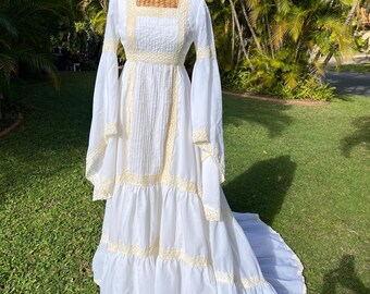 May include: A floor-length white dress featuring long bell sleeves, a square neckline, and vertical pleats on the bodice. The garment is accented with cream-colored lace trim along the neckline, waist, sleeves, and tiered skirt hemline.