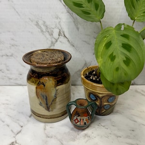Könnte beinhalten: Ein Keramiktopf mit Korkdeckel, in Braun- und Beigetönen mit einem blattartigen Design. Daneben eine kleine dekorative Vase mit geometrischem Muster und grünem Griff. Eine Topfpflanze mit grünen Blättern ist ebenfalls vorhanden.