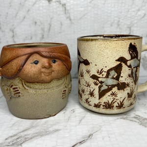 May include: Two ceramic mugs. One is a tan mug with a handle and a design of flying ducks. The other is a tan and brown mug with a face design.