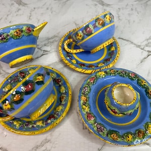 Puede incluir: Un juego de cuatro tazas y platillos de cerámica azul. Las tazas y los platillos tienen rayas amarillas y están decorados con diseños florales y frutales coloridos. Las tazas tienen asas y los platillos tienen un diseño a juego. El juego está sobre una superficie de mármol blanco.