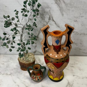 May include: A decorative arrangement featuring a small, handcrafted tree with green gemstone leaves, a small ceramic vase with a green handle, and a larger terracotta vase with a detailed painted design. The vases have a rustic, artisanal aesthetic.