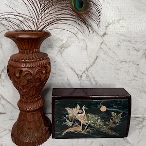 Può includere: Un vaso di legno intagliato con una piuma di pavone e una scatola decorativa. Il vaso è marrone scuro con intagli intricati. La scatola è marrone scuro con un disegno intarsiato verde e oro di uccelli e fogliame.