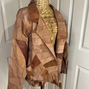 May include: A brown and tan patchwork leather jacket. The jacket has a collared neckline, two front pockets, and a tie belt. The design is made of various geometric shapes.