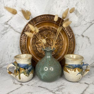 May include: Two ceramic mugs with a speckled cream base and blue and brown drip glaze patterns, flanking a round, sage green ceramic vase containing dried tan bunny tail grass, all positioned in front of a large, round, carved wooden plate.