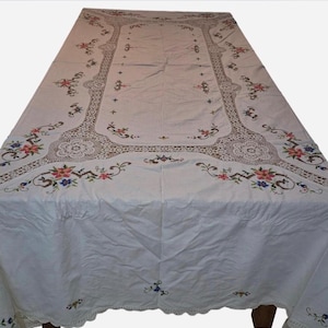 Pode incluir: Uma toalha de mesa retangular branca com inserções de renda intrincadas e bordados florais coloridos. O design inclui delicados padrões de costura de pequenas flores em tons de vermelho, azul e verde, dispostos ao longo das bordas.