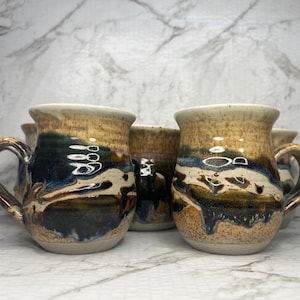 May include: A set of four ceramic mugs featuring a rounded shape and a handle on the side. The mugs have a speckled tan glaze on the upper portion, transitioning into a dark blue and brown drip pattern across the middle and lower sections.
