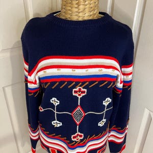 May include: A navy blue sweater with a crew neck and long sleeves. The sweater features red, white, and tan stripes, along with embroidered geometric patterns. The cuffs and hem are trimmed in red.