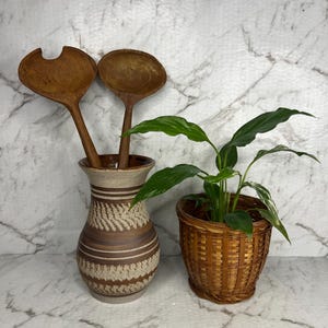 May include: A brown ceramic vase with a textured pattern holds two wooden salad spoons. A small potted plant in a woven basket sits to the right. The background is a white marble pattern.