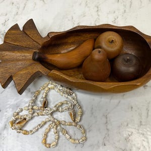 May include: A carved wooden pineapple-shaped bowl containing three wooden fruit figures: a banana, a pear, and an apple. A necklace made of small, light-coloured seashells is positioned below the bowl. The wood has a rich, medium-brown grain.