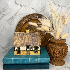 May include: A ceramic cottage figurine with a textured brown thatched roof, cream-colored walls, and a yellow base, resting on two stacked teal books next to a carved wooden vase filled with dried beige grasses and a round wooden plaque.