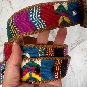 Op de afbeelding: Een kleurrijke geweven riem met een bruine leren achterkant en afwerking. De riem heeft een levendig patchwork-ontwerp met rode, blauwe, paarse en gele geometrische patronen. De riem heeft een metalen gesp en oogjes.