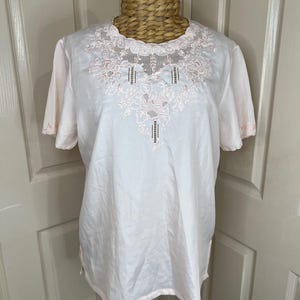May include: A pale pink short-sleeved top with detailed floral embroidery around the neckline and centre. The embroidery uses delicate pink thread. The top is displayed on a mannequin.
