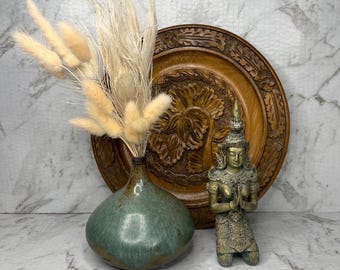 Könnte beinhalten: Eine runde, blaugrün glasierte Keramikvase mit getrockneten beigen Grasbüscheln, neben einer kleinen, knienden Metallfigur mit gefalteten Händen, vor einem großen, runden, geschnitzten dekorativen Holzteller platziert.