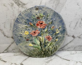 May include: A round, ceramic wall hanging with a floral design. The background is a textured blue, and the flowers are red, yellow, and green. The piece has a hole at the top for hanging.