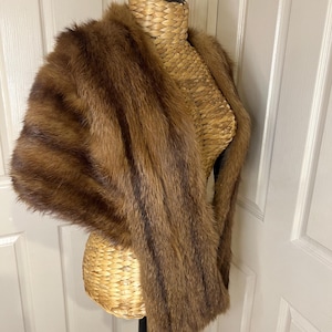 May include: A brown fur stole with dark brown stripes is draped over a wicker mannequin. The stole is displayed against a white door background. The fur appears soft and luxurious, with a natural, textured appearance.