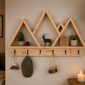 Peut inclure: Une étagère murale en bois clair en forme de sommets montagneux avec six crochets en métal noir fixés en bas. L'étagère présente un petit arbre artificiel, une pomme de pin, une figurine de cerf noir et un panneau indiquant EXPLORE DREAM DISCOVER.