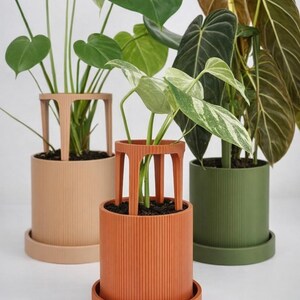 Peut inclure: Trois pots de plantes décoratifs beiges, oranges et verts. Chaque pot est accompagné d'une soucoupe assortie et d'un support unique. Les pots sont remplis de diverses plantes vertes, mettant en valeur un design moderne pour la décoration intérieure.