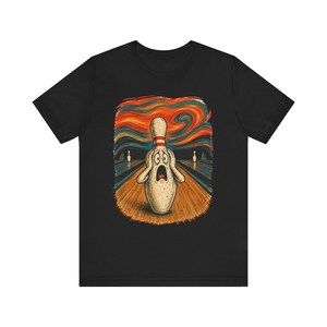 Può includere: Una t-shirt nera con una stampa grafica di un birillo da bowling bianco con un'espressione scioccata, le mani sul viso, in piedi su una pista di legno con uno sfondo di cielo arancione e blu vorticoso e due birilli in lontananza.