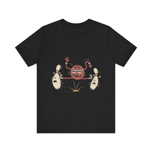 Può includere: Una t-shirt nera a maniche corte con un'illustrazione grafica di una palla da bowling rossa con braccia e gambe che salta sopra un piccolo schizzo giallo, affiancata da due birilli bianchi con espressioni facciali sorprese.