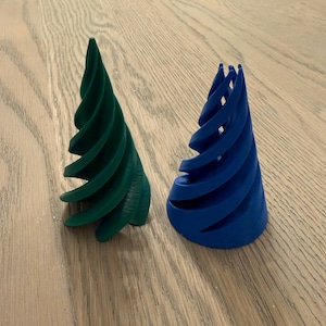 以下が含まれることがあります： 木製の表面に置かれた、緑と青の2つの3Dプリントされたらせん状の円錐形のツリー