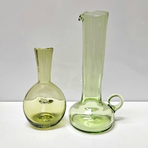 May include: Two green glass vases. One is a round bottle shape with a narrow neck. The other is a tall, slender vase with a handle and a spout. Both vases have a light green tint and are transparent.