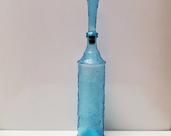May include: A tall, light blue glass bottle with a long, decorative stopper. The bottle features an embossed floral pattern and a cork. The stopper is also blue and has a textured design. The bottle is approximately 12 inches tall.
