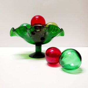 May include: A green glass bowl with a ruffled edge and a pedestal base. The bowl is filled with red, green, and yellow glass balls.